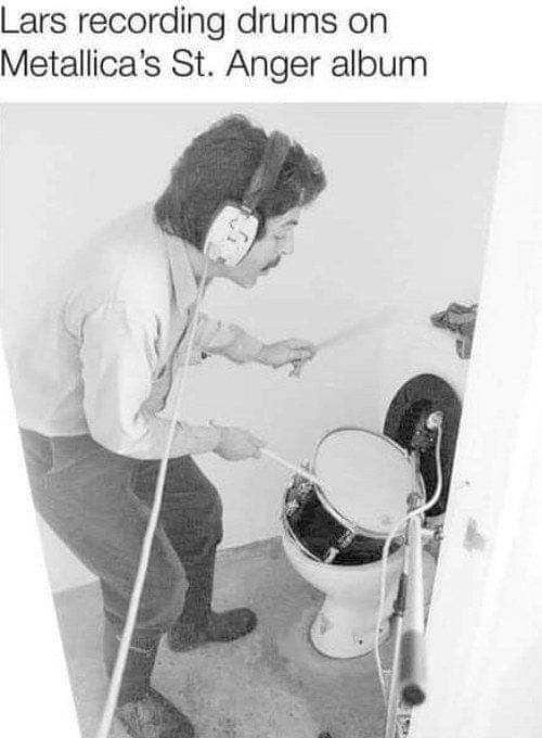 A black-and-white photo shows a man with headphones and drumsticks playing a snare drum that is mounted on a toilet bowl…