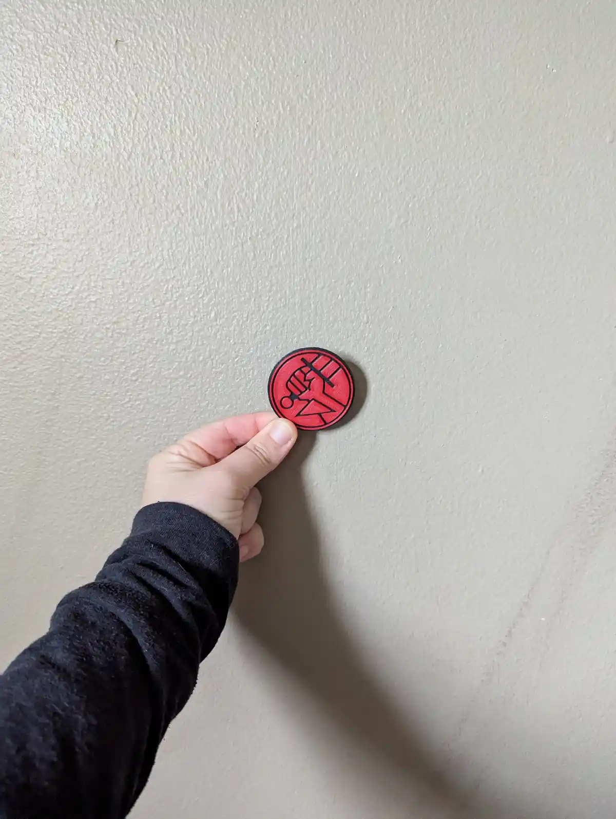 A hand holds a small circular red and black pin against a plain beige wall. The pin features a stylized fist logo. Overlaid…