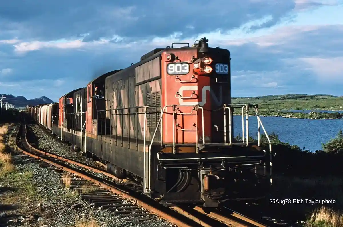 A vintage red and black Canadian National Railway locomotive, numbered 903, pulls a long freight train along a scenic track…