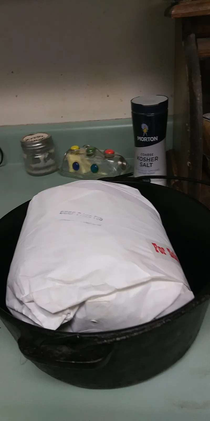 A large white paper-wrapped package labeled 'beef prime rib' sits inside a black cast iron pot on a green countertop. In the…