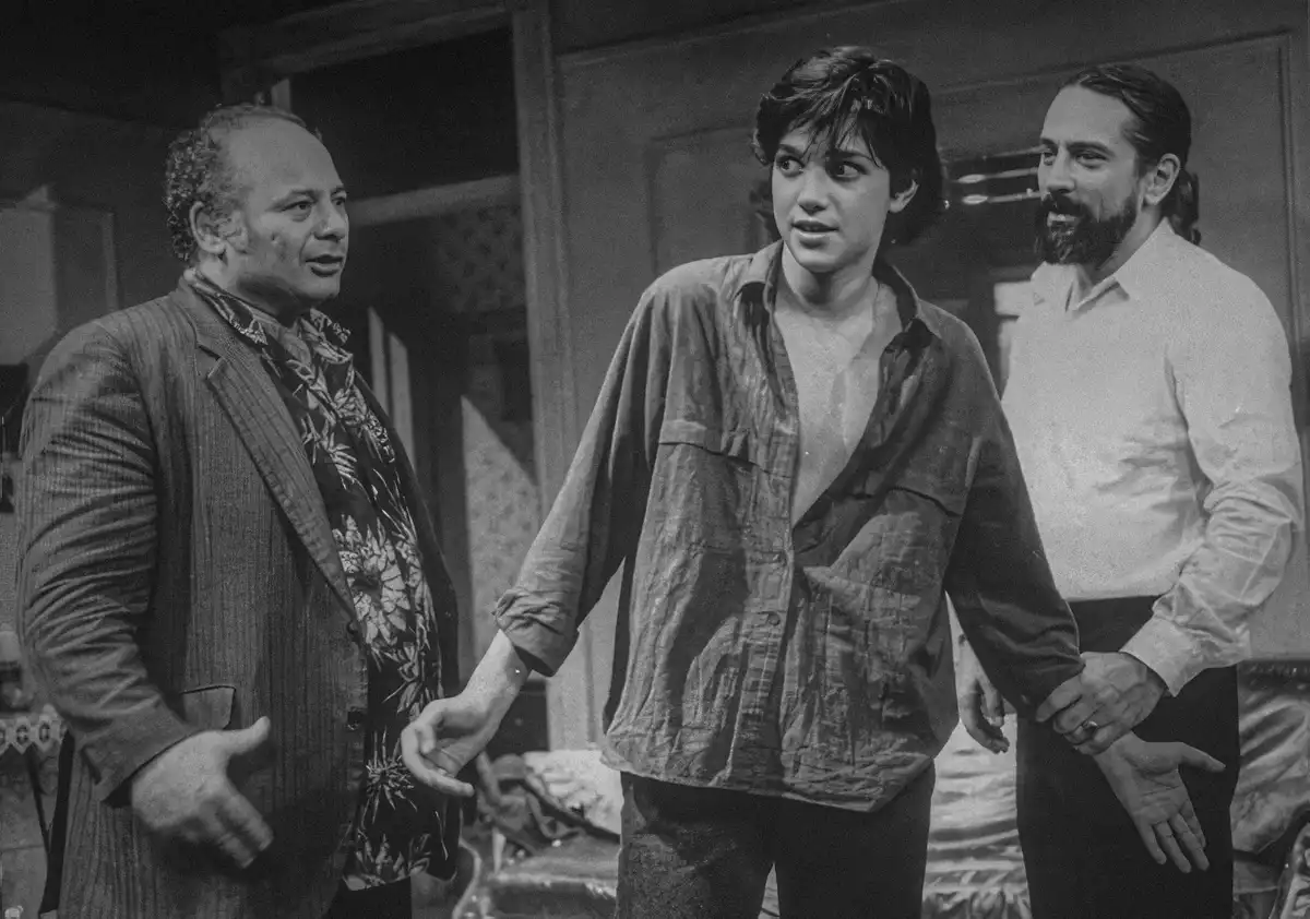 A black-and-white photo shows three men standing together indoors. The man on the left wears a patterned shirt under a…