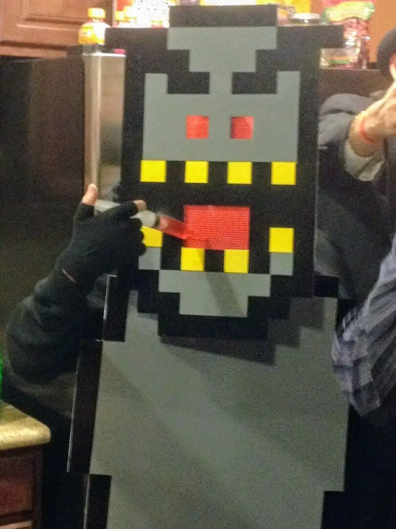 A person in a large, pixelated, gray and black costume resembling a classic video game monster holds a syringe filled with…