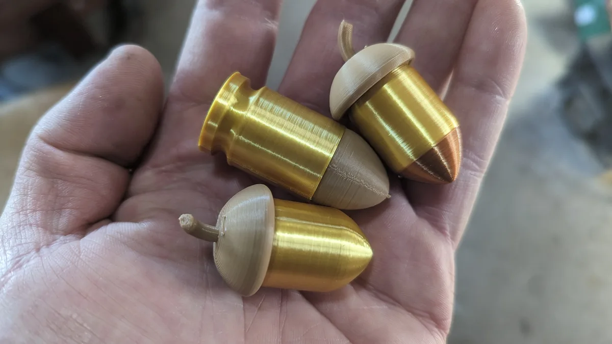 A person's open palm holds three 3D-printed objects shaped like acorns, with gold-colored bodies and beige caps and stems…