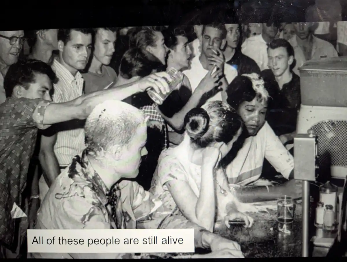 A black-and-white photo shows a crowded, vintage-looking bar scene with many people gathered around a counter. A man in a…
