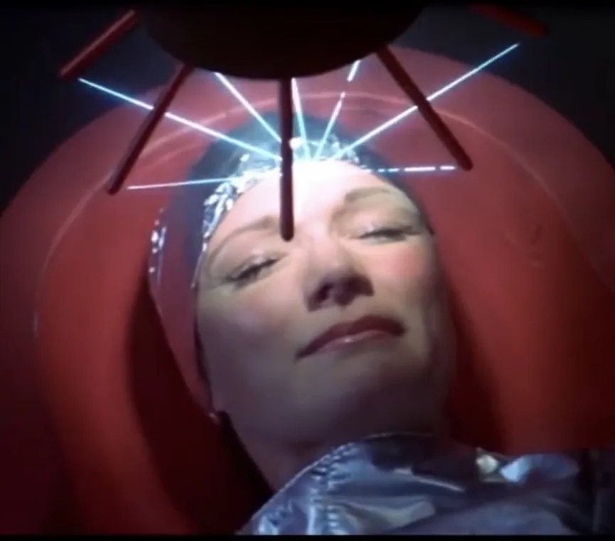 A woman with foil on her head lies in a red chair as blue laser beams shoot from a device above her head. The overlaid text…