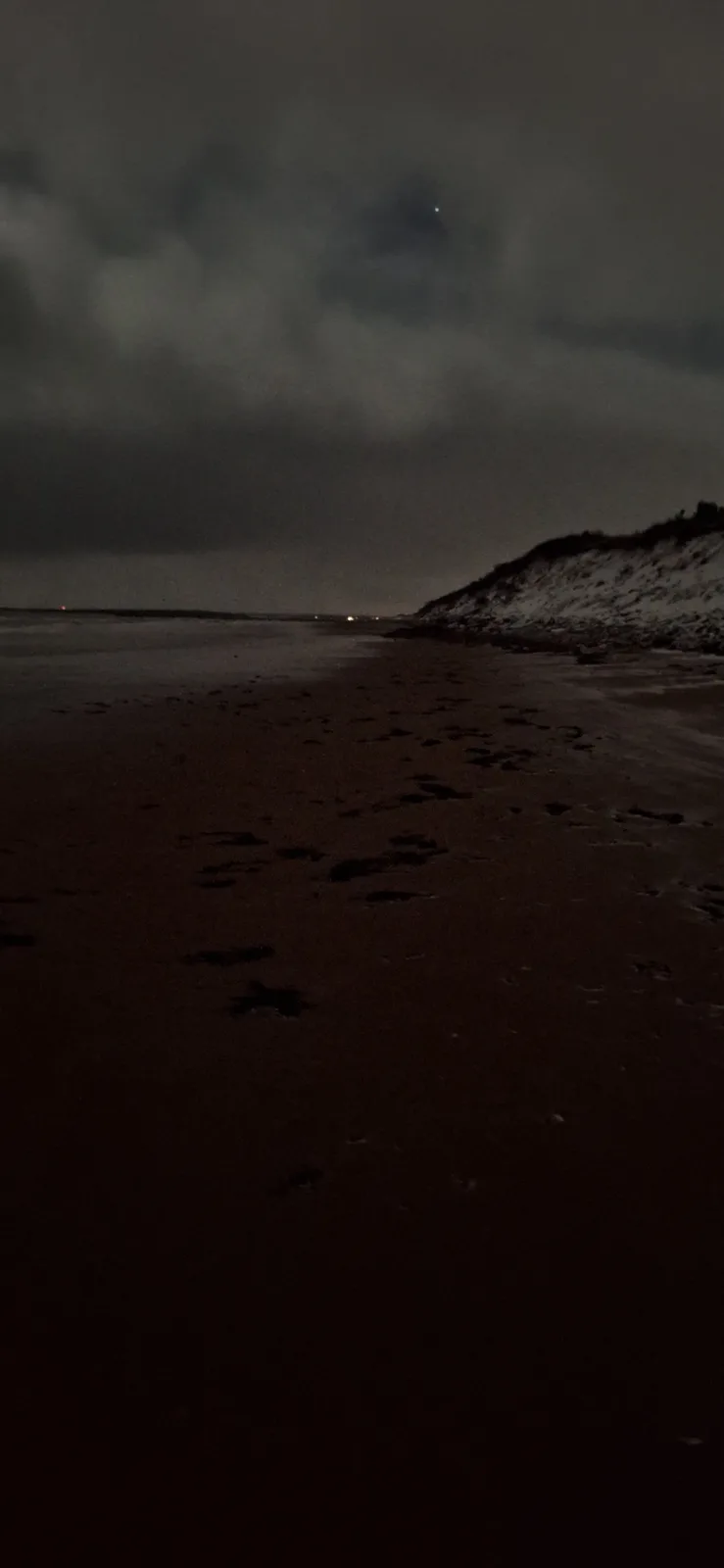A dark, low-light photo of a beach at night with footprints in the sand leading toward the water, a snowy dune on the right…