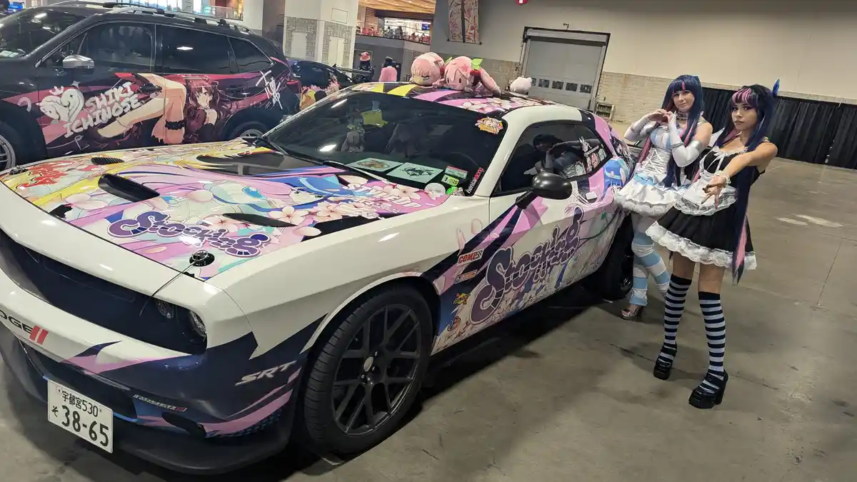 A white Dodge Challenger with a vibrant anime-style wrap featuring the word 'Stocking' and cherry blossoms is parked…