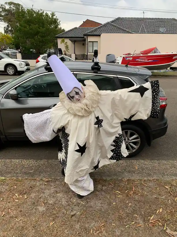 A person dressed as a clown with a tall purple hat, white face paint with black accents, and a large ruffled collar stands…