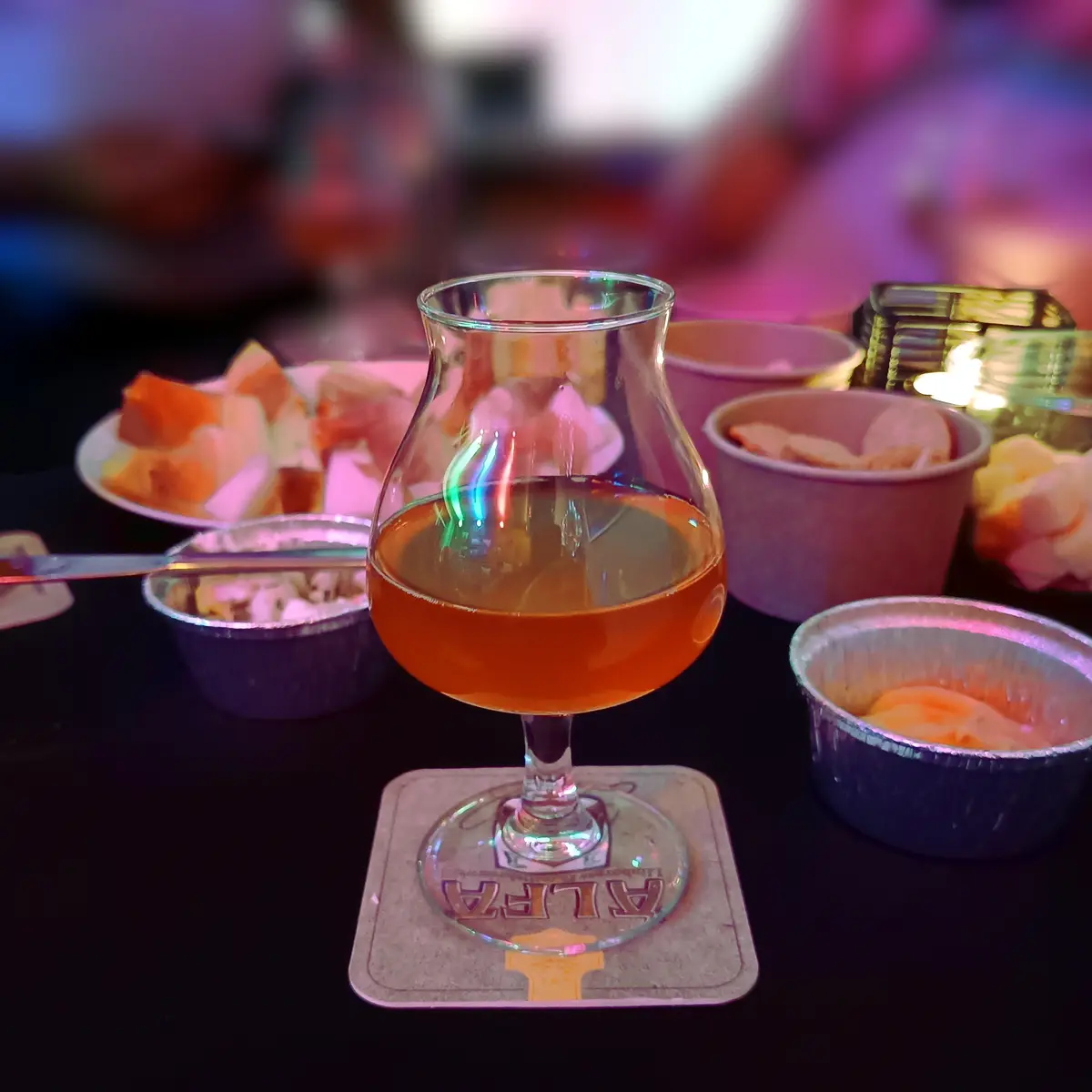 A close-up photo shows a stemmed glass filled with amber liquid, likely beer or whiskey, resting on a coaster with 'ALFA'…