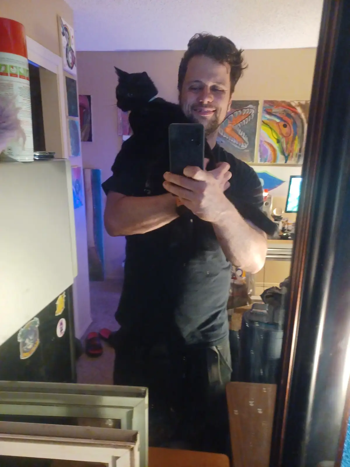 A man with dark hair and a beard takes a mirror selfie while holding a black cat on his shoulder. He smiles at the phone…
