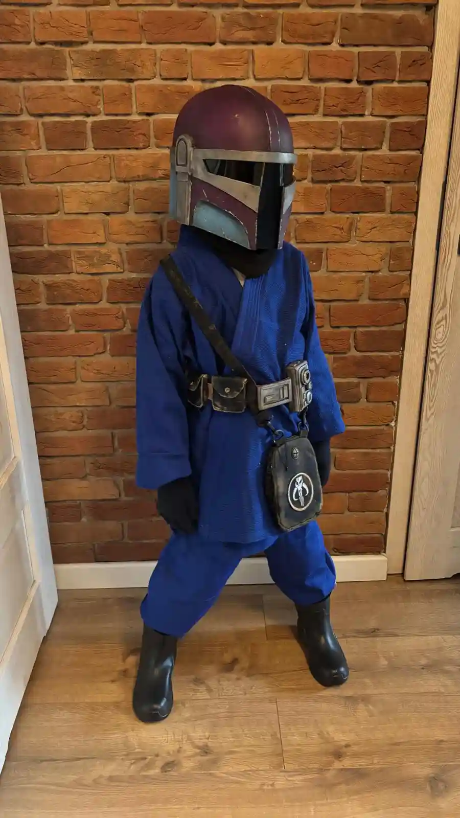 A child stands in front of a brick wall wearing a Mandalorian helmet, blue Jedi robes, black gloves, and black boots. The…