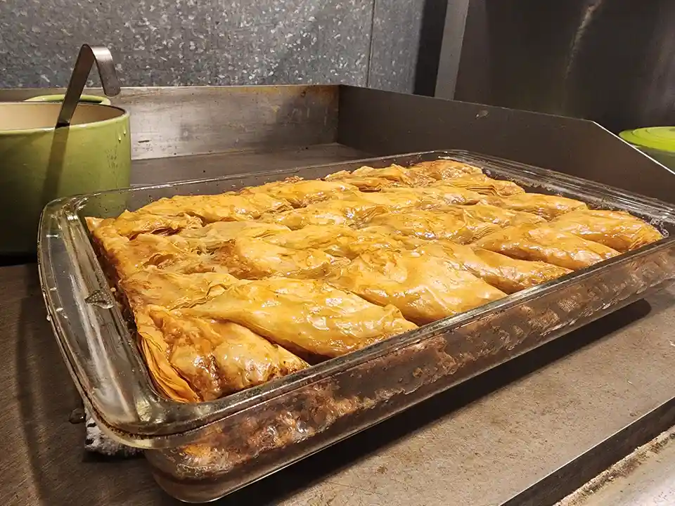 A close-up photo shows a glass baking dish filled with golden-brown, flaky baklava, glistening with syrup, sitting on a…