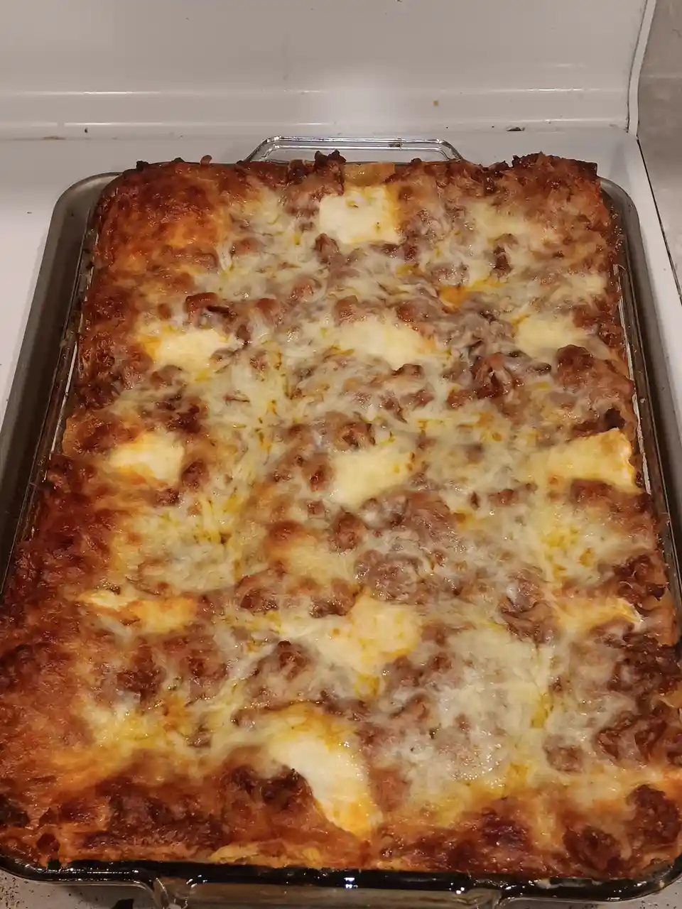 A freshly baked lasagna in a glass baking dish sits on a white stovetop, its surface covered in melted, bubbly cheese with…