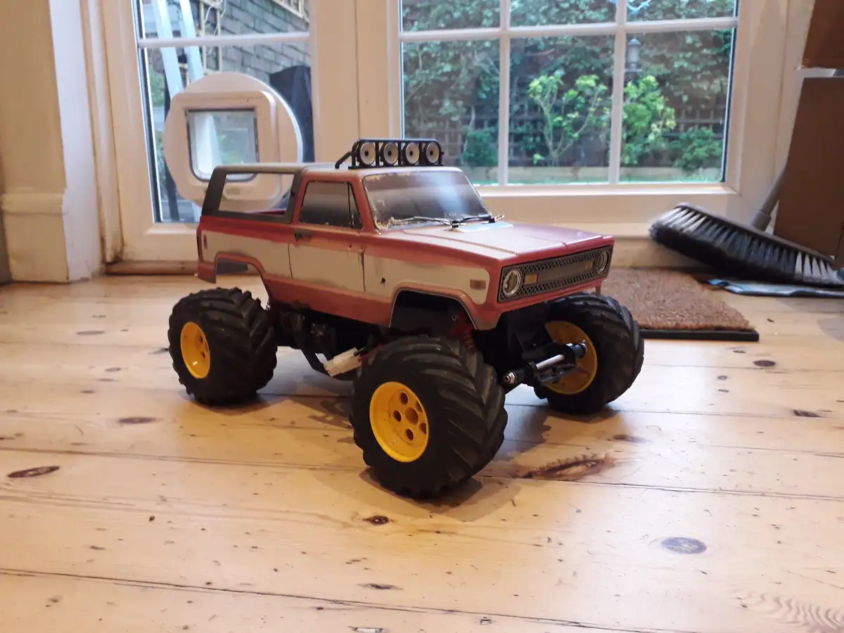 A red and white remote-controlled monster truck with oversized black tires and yellow rims sits on a light wooden floor…