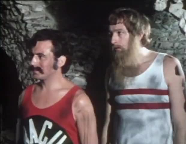 Two men in vintage tank tops stand in a dimly lit cave-like setting. The man on the left wears a red tank top with a…