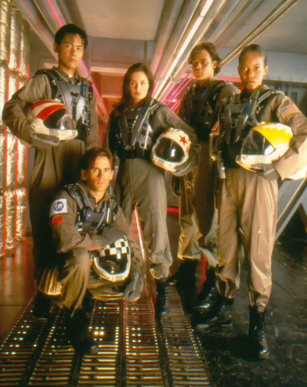Five people in space flight suits pose in a spaceship corridor, each holding a helmet. The person in the front, kneeling…