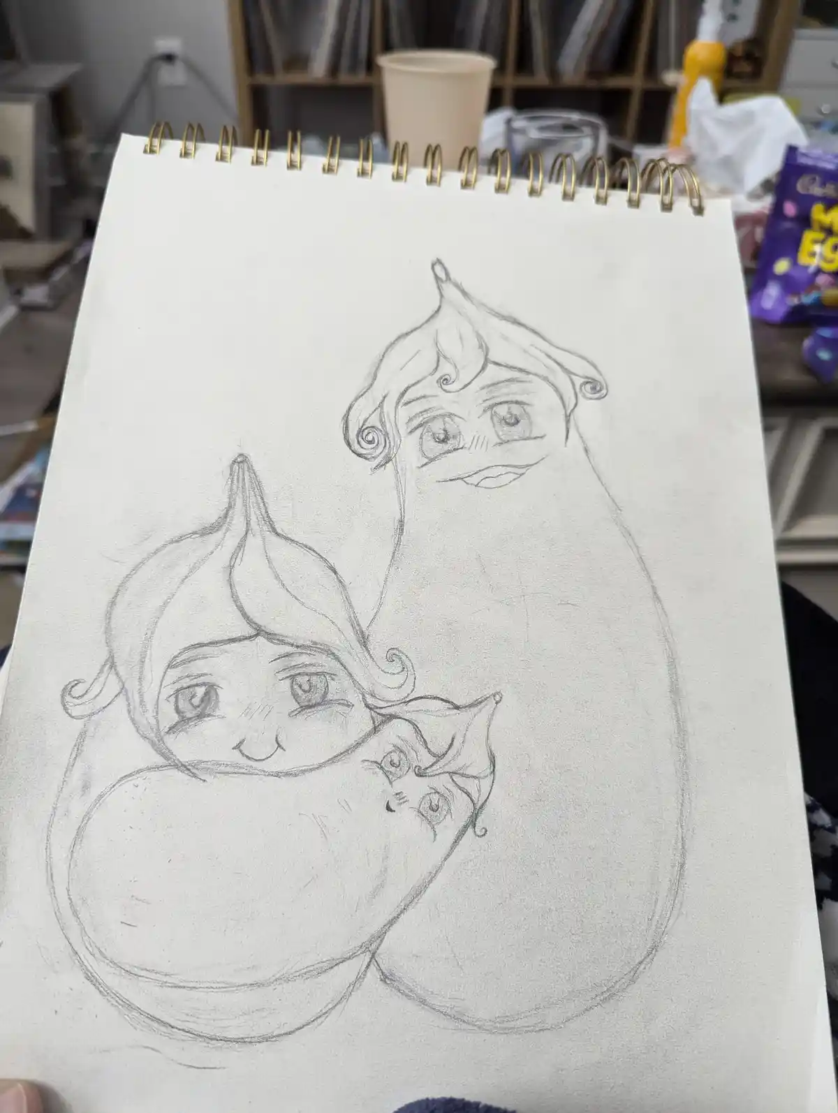 A pencil sketch on a spiral-bound notebook shows three anthropomorphic eggplants with cartoonish faces, each with a smiling…