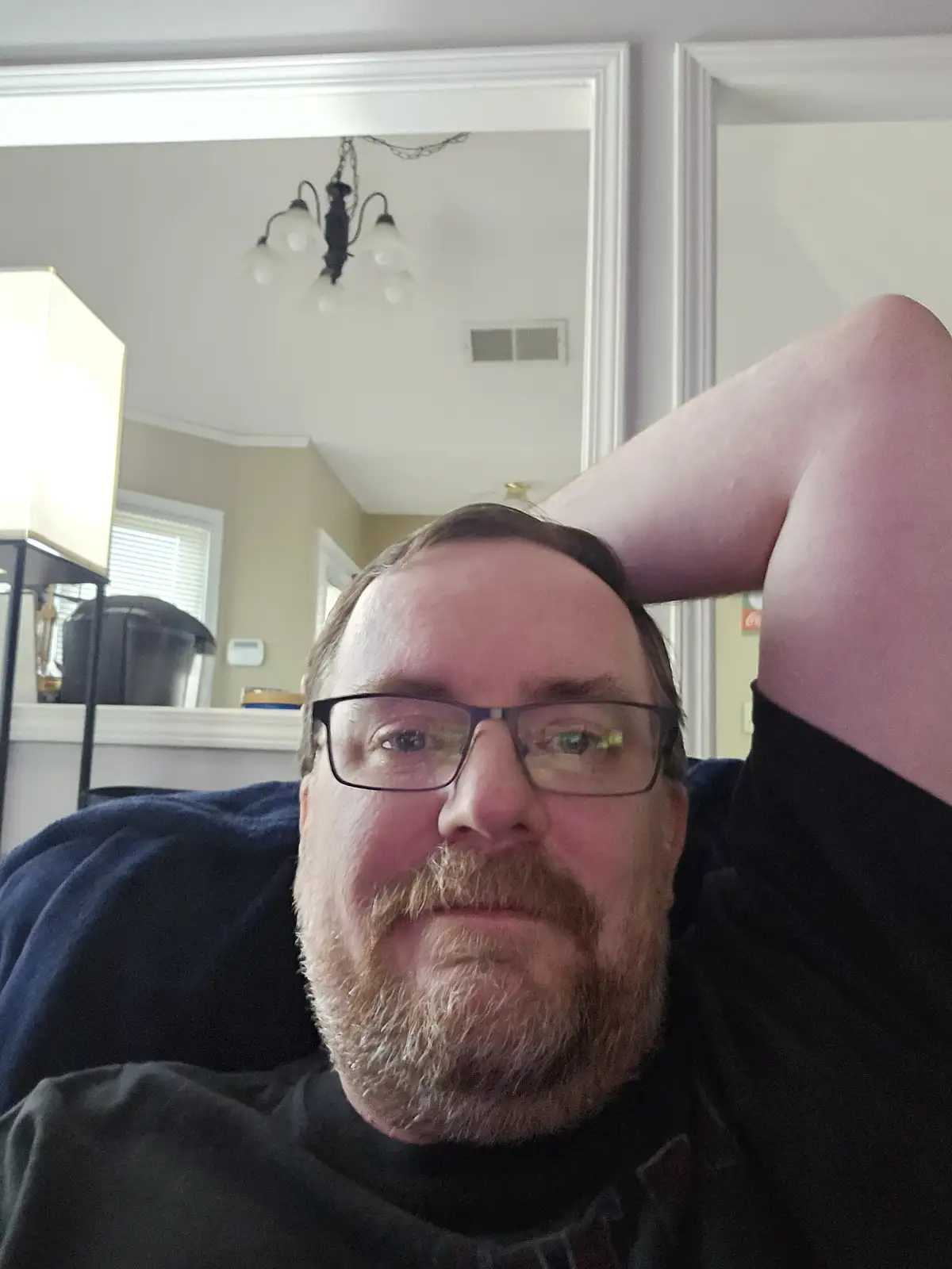 A man with glasses and a beard, wearing a black shirt, lies down with his arm behind his head in a relaxed pose. The…