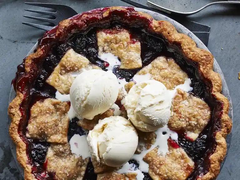 An overhead shot of a rustic berry pie with a golden-brown, fluted crust, topped with three scoops of vanilla ice cream…