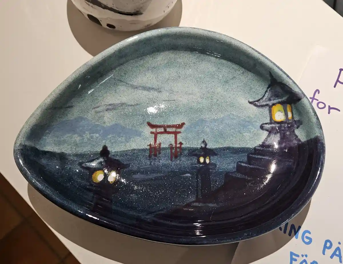 A ceramic plate with a painted scene of a Japanese torii gate and lanterns, where the lanterns have cartoonish glowing eyes…