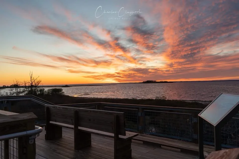 A serene sunset photograph shows a wooden deck with benches overlooking a calm body of water, with vibrant orange and pink…