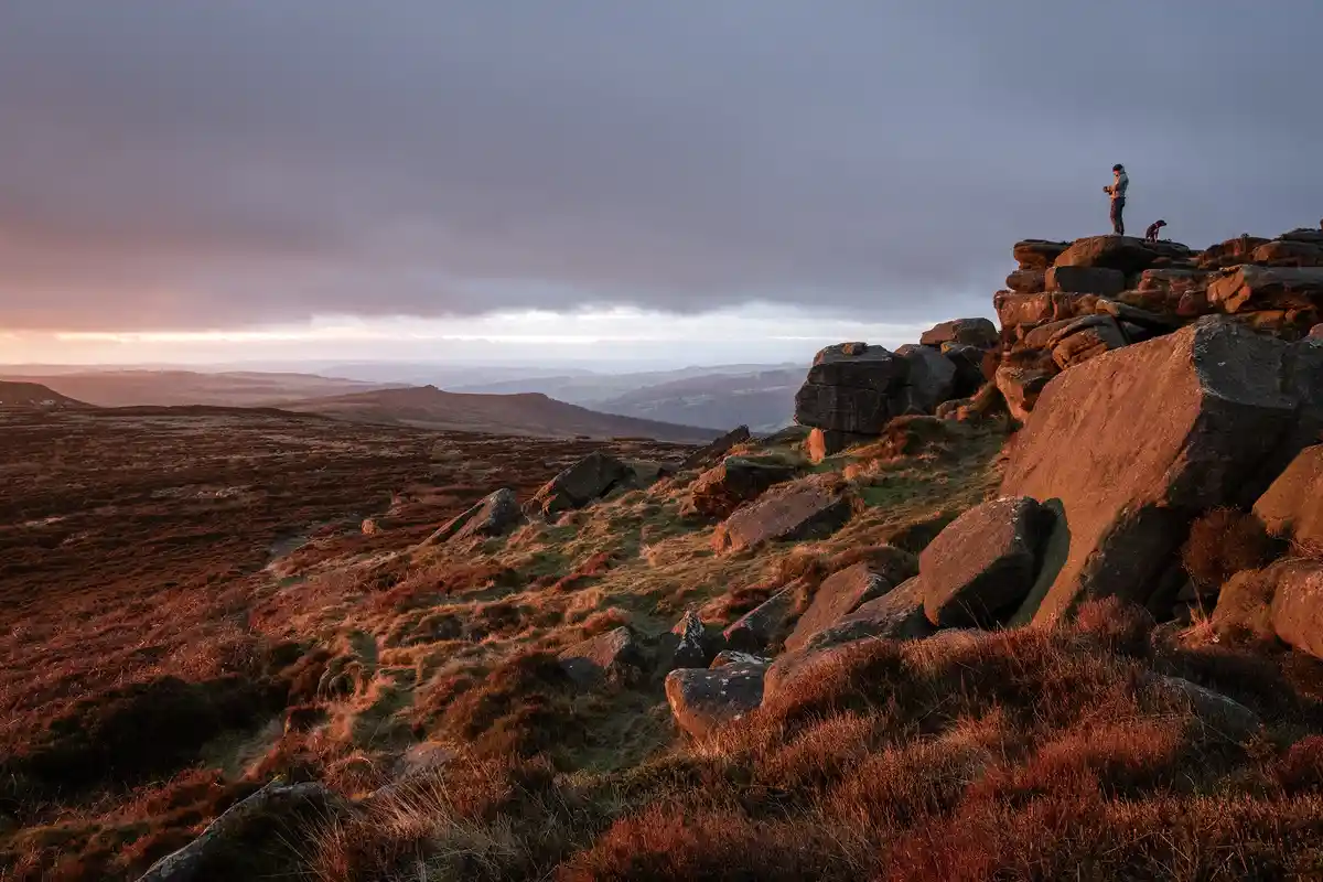 A person and a dog stand atop a rocky hill during a dramatic sunset, overlooking a vast, hilly landscape. The person is…