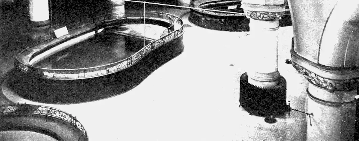A black-and-white photo shows an ornate, curved, empty pool or basin surrounded by decorative pillars and railings, with…