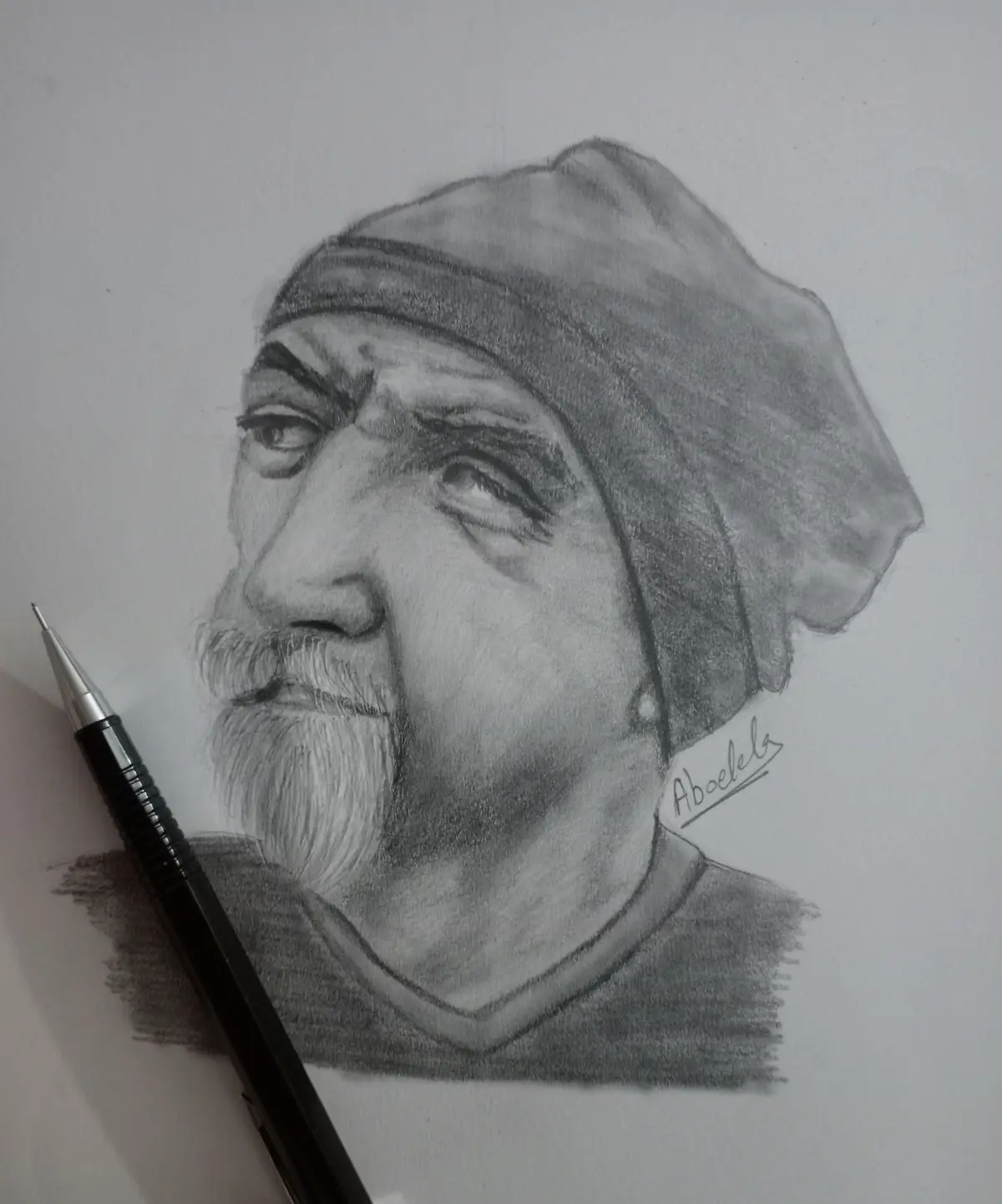 A detailed pencil sketch of a man with a beard and head covering, rendered in grayscale, with a black pen resting on the…