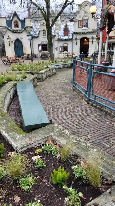 A green wooden bench is awkwardly placed at an odd angle inside a stone garden bed in a European-style theme park courtyard…