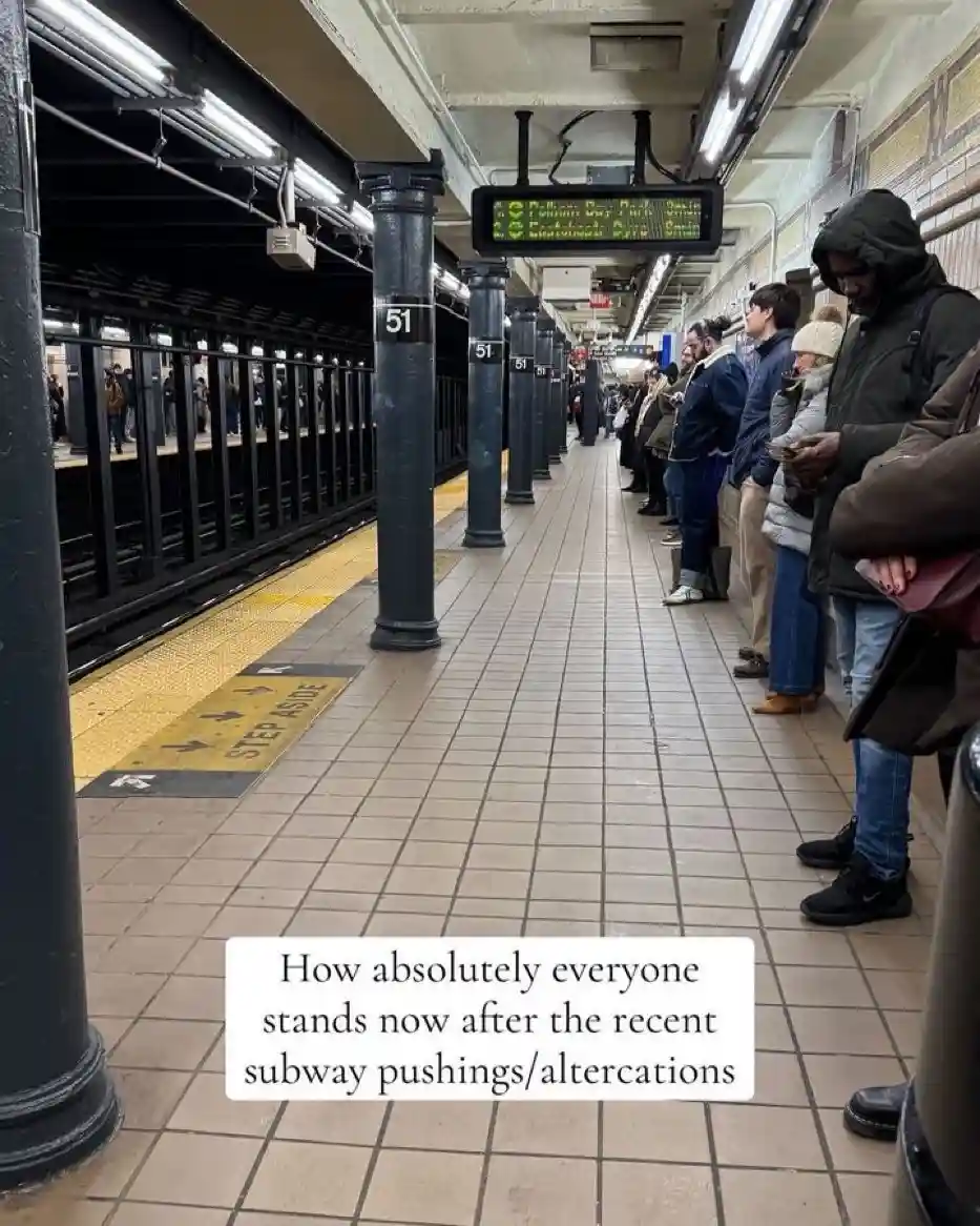A subway platform with people standing in a single-file line, facing away from the camera, near a train. The platform has…