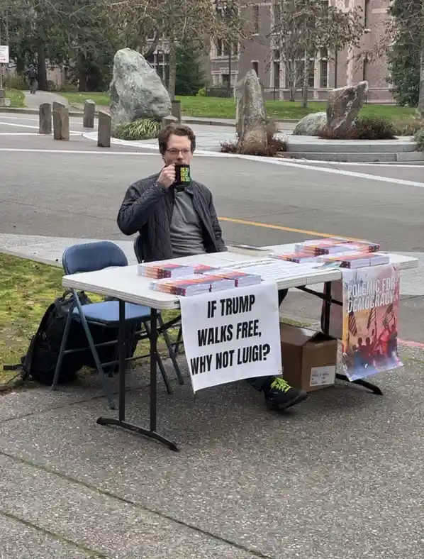 A man sits at a folding table outdoors, holding a mug, with stacks of books and a banner reading 'IF TRUMP WALKS FREE, WHY…