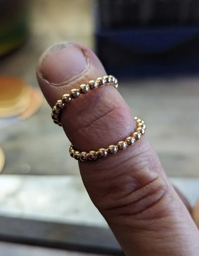 A close-up photo shows two identical gold bead rings worn on a single finger, one stacked above the other. The rings are…