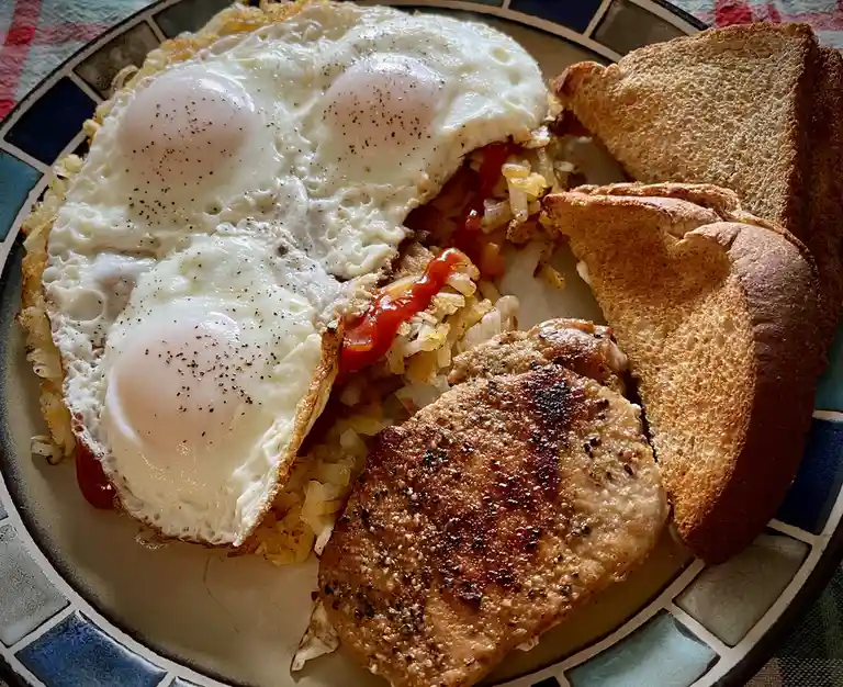A close-up photo of a hearty breakfast plate featuring three sunny-side-up eggs seasoned with black pepper, crispy hash…