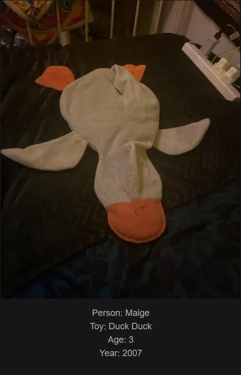 A close-up photo of a worn, handmade fabric duck toy lying on a dark patterned bedspread. The toy has a light gray body…