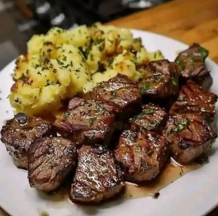 A close-up photo of a white plate holding a generous serving of seared, juicy steak cubes with visible grill marks and a…