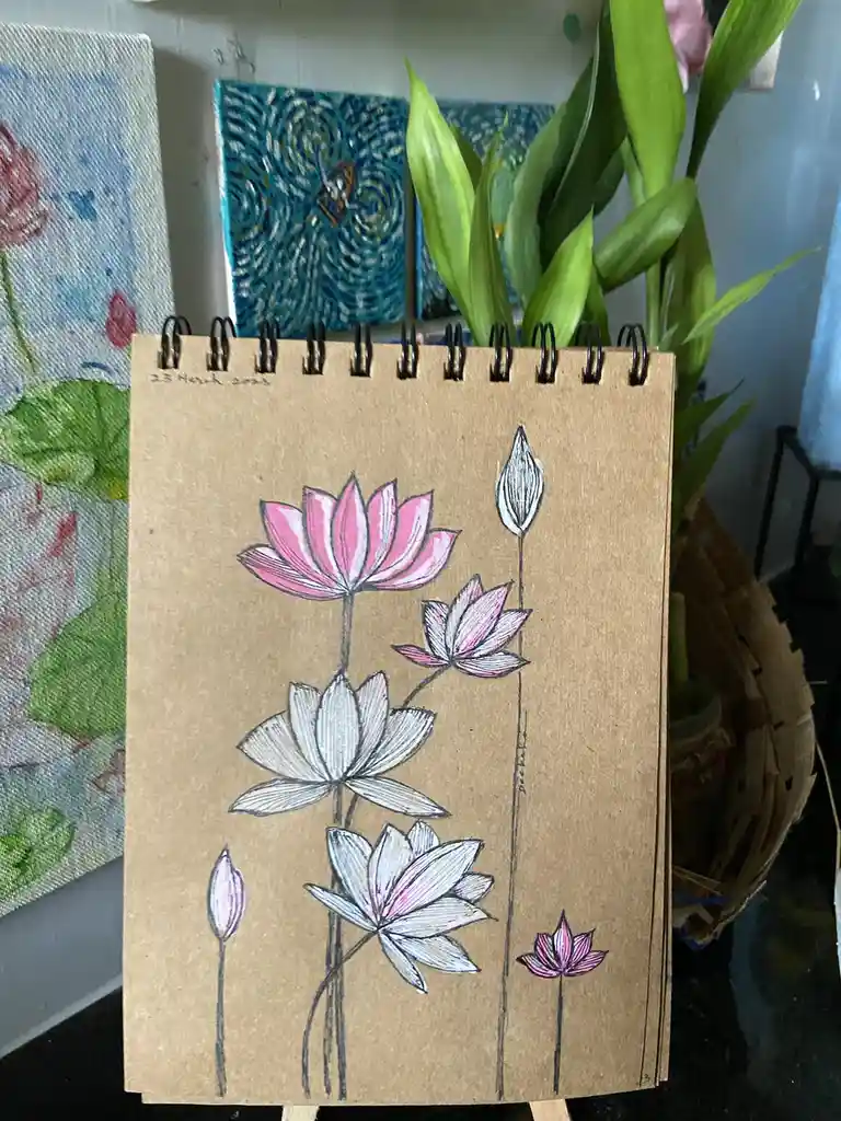A spiral-bound sketchbook with brown kraft paper pages displays a hand-drawn illustration of several lotus flowers in…
