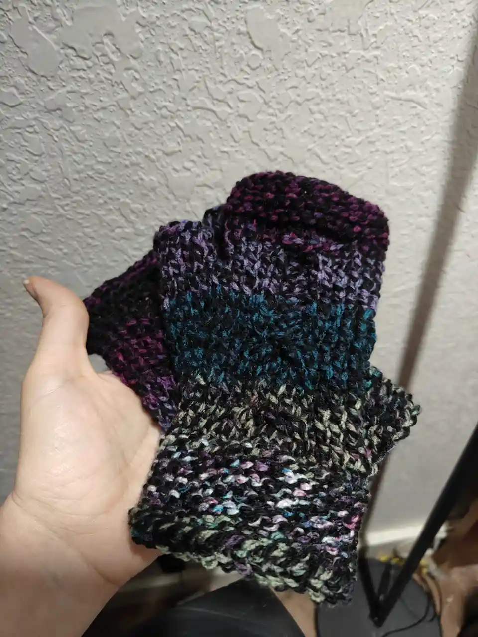 A hand holds up a hand-knitted scarf with a gradient of dark colors, starting with purple and black at the top…
