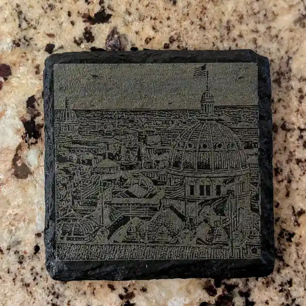 A square black stone coaster with an engraved, detailed cityscape featuring a prominent domed building topped with an…