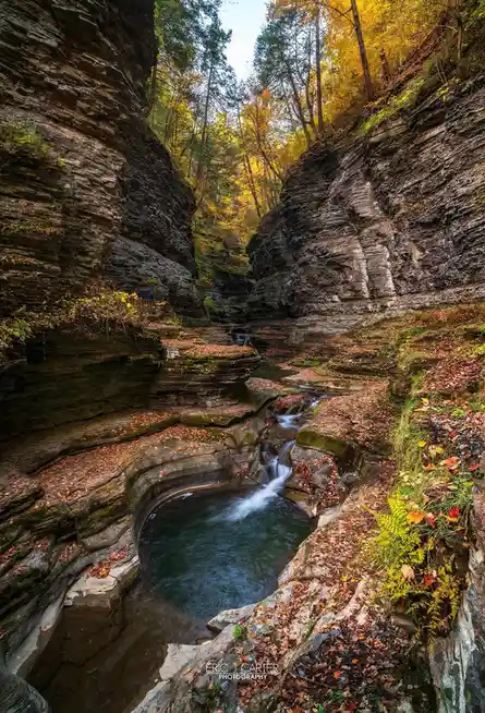 A scenic autumn photograph of a narrow rocky gorge with a small waterfall flowing into a pool, surrounded by trees with…