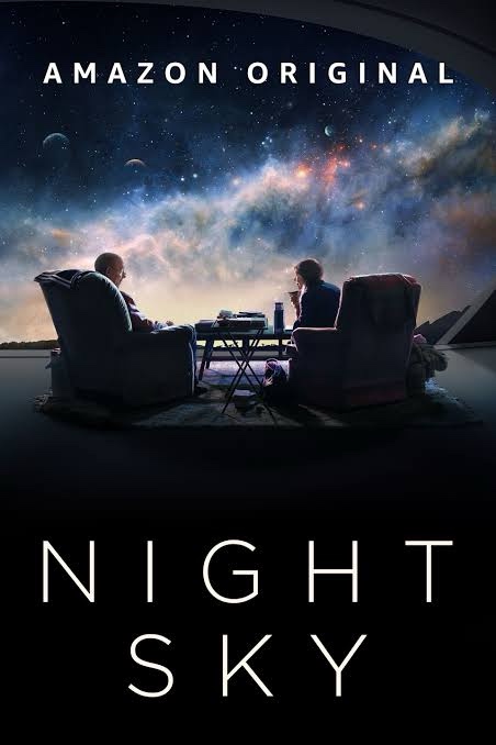 Two people sit in armchairs facing each other across a small table, gazing out at a vast, star-filled galaxy with planets…