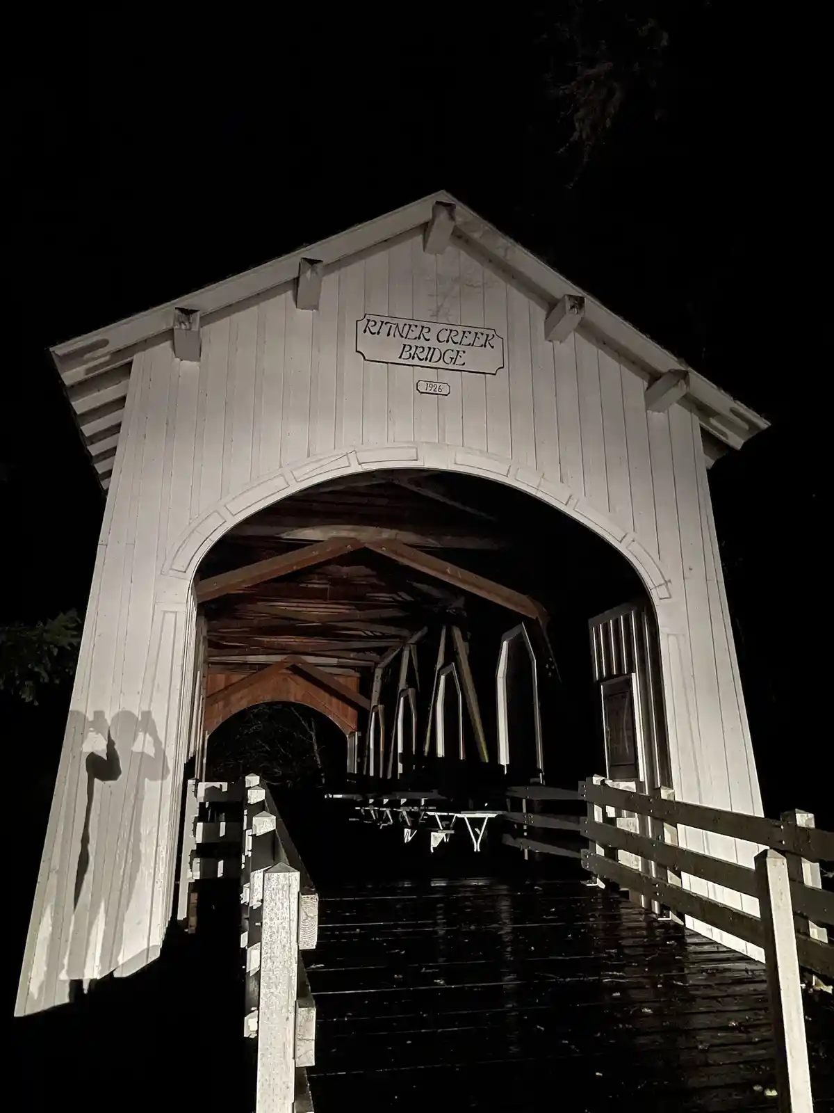 A low-angle nighttime photo shows the entrance to a white wooden covered bridge labeled 'RITNER CREEK BRIDGE 1926'. The…