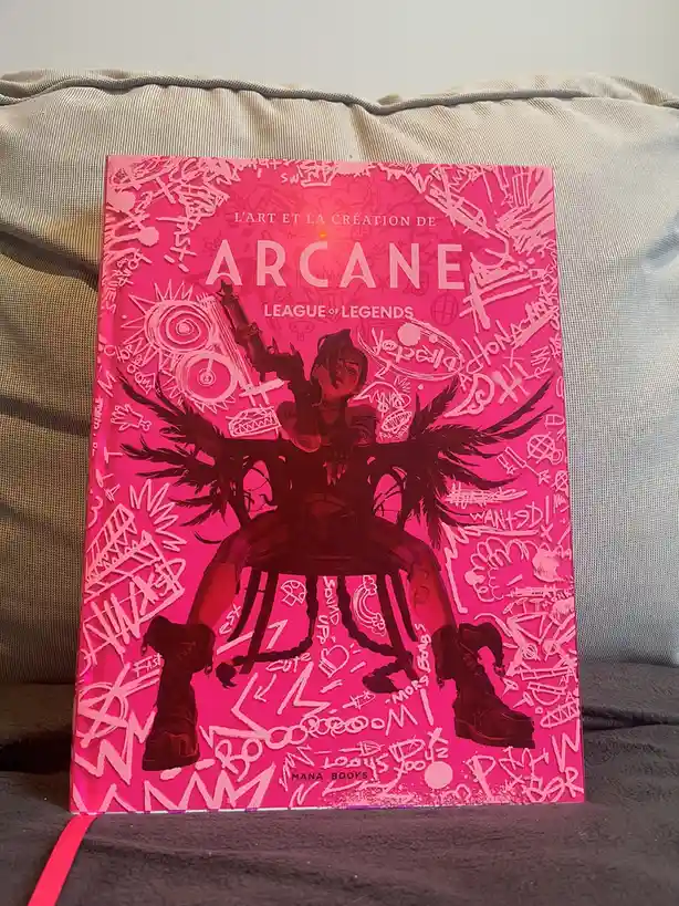 A bright pink hardcover art book titled 'L'ART ET LA CRÉATION DE ARCANE LEAGUE OF LEGENDS' rests on a beige couch. The cover…