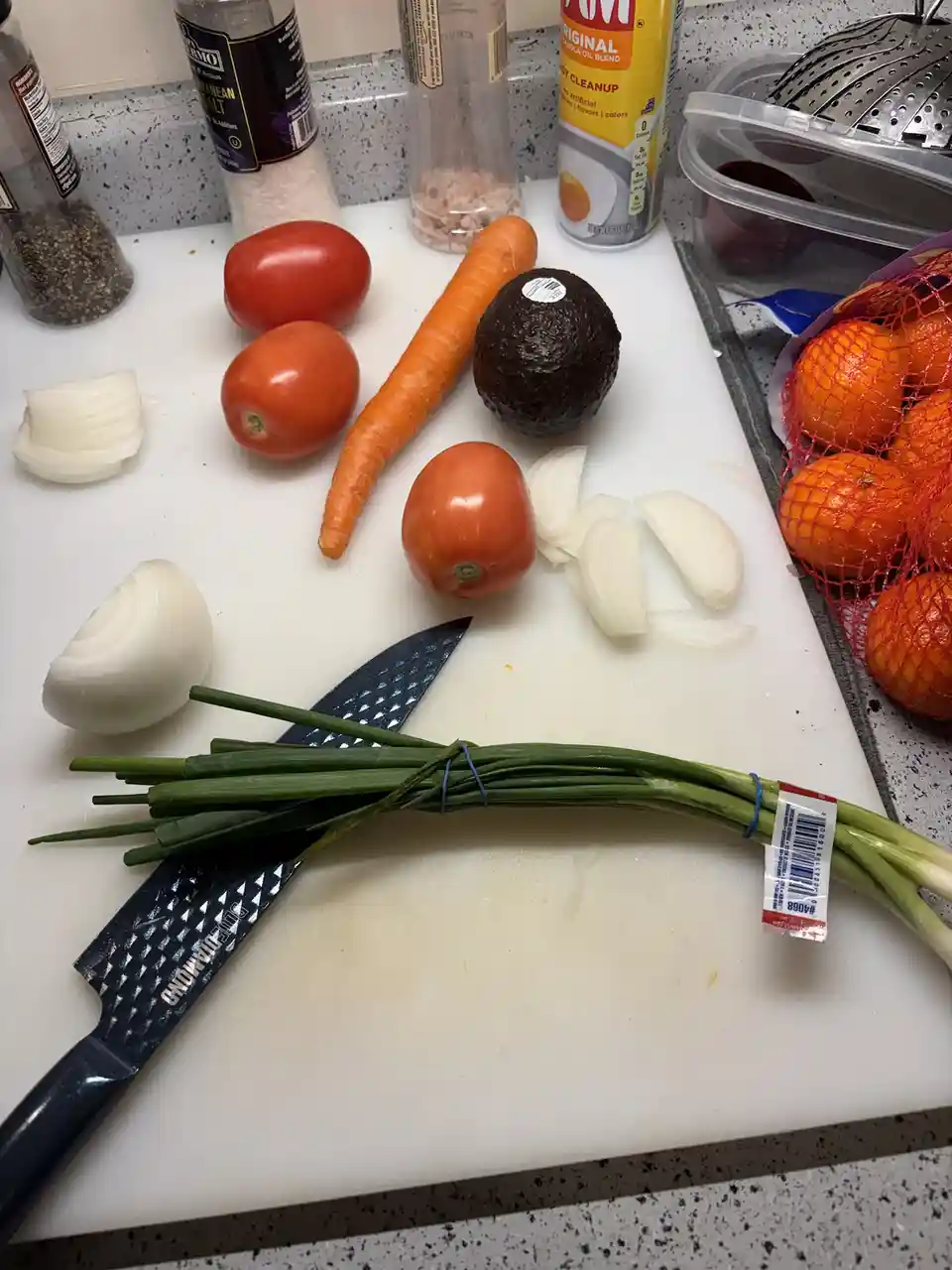 A white cutting board holds chopped onions, whole tomatoes, a carrot, an avocado, and a bundle of green onions next to a…