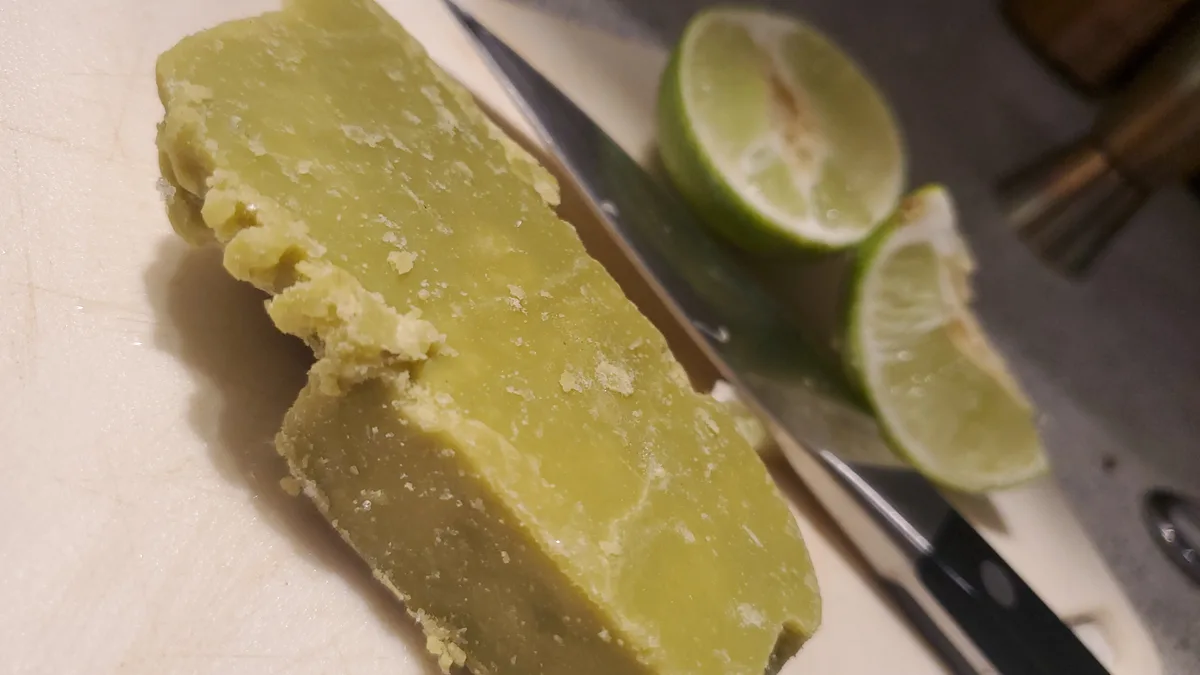 A close-up photo shows a large, pale green, crumbly block of what appears to be lime zest or lime paste on a white cutting…