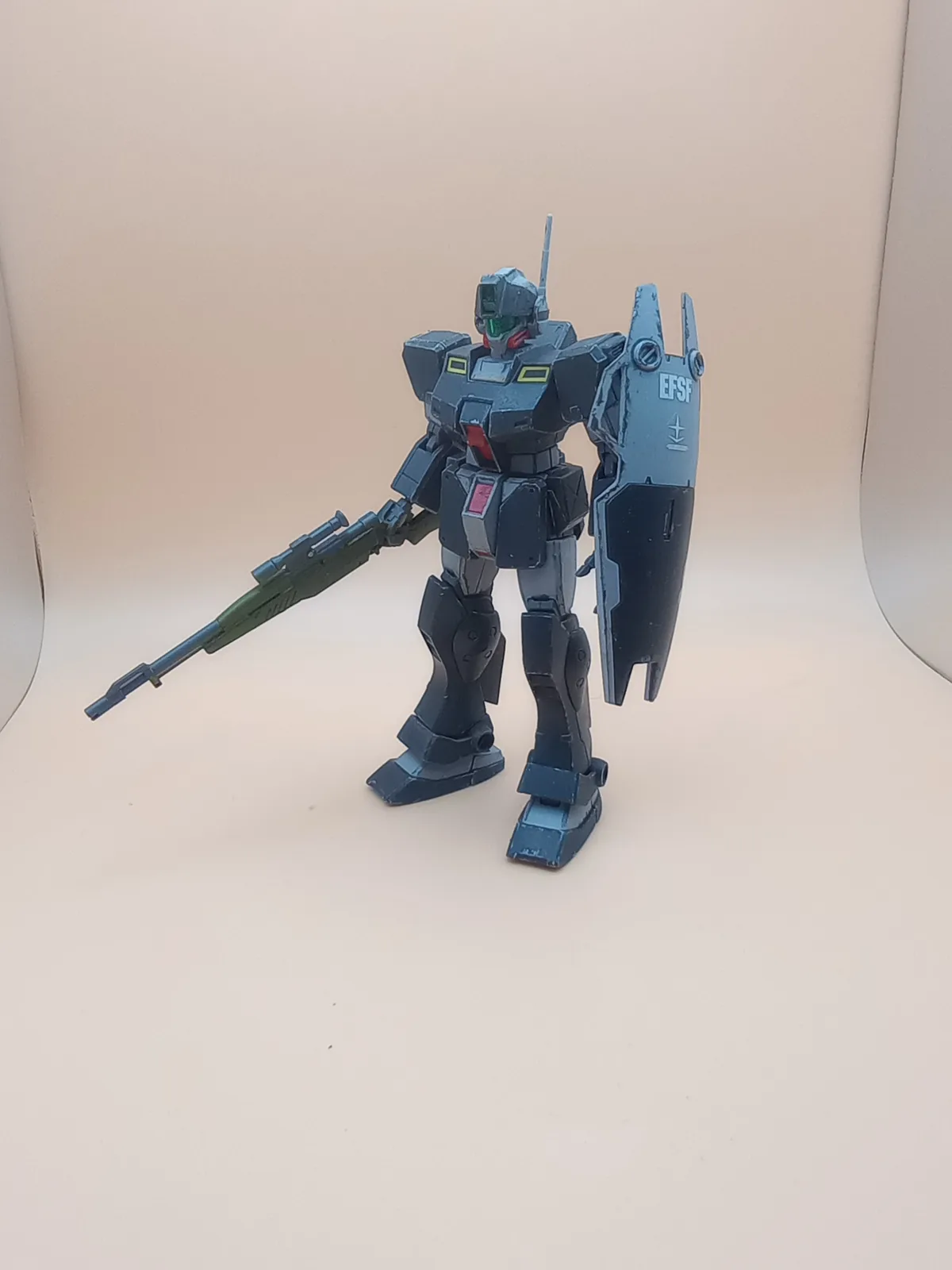 A dark grey, weathered mobile suit model stands against a plain beige background, holding a rifle in its left hand and a…
