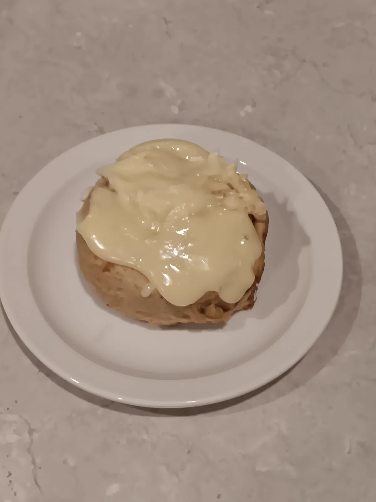 A single cinnamon roll sits on a white plate, generously covered in a thick, creamy white frosting that drips slightly over…