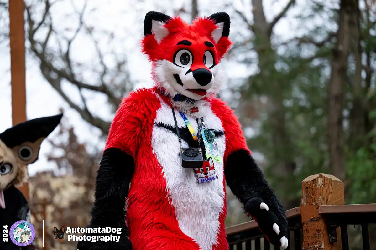 A person in a red and white fox fursuit stands outdoors, wearing a small black fan clipped to their chest. Another fursuit…