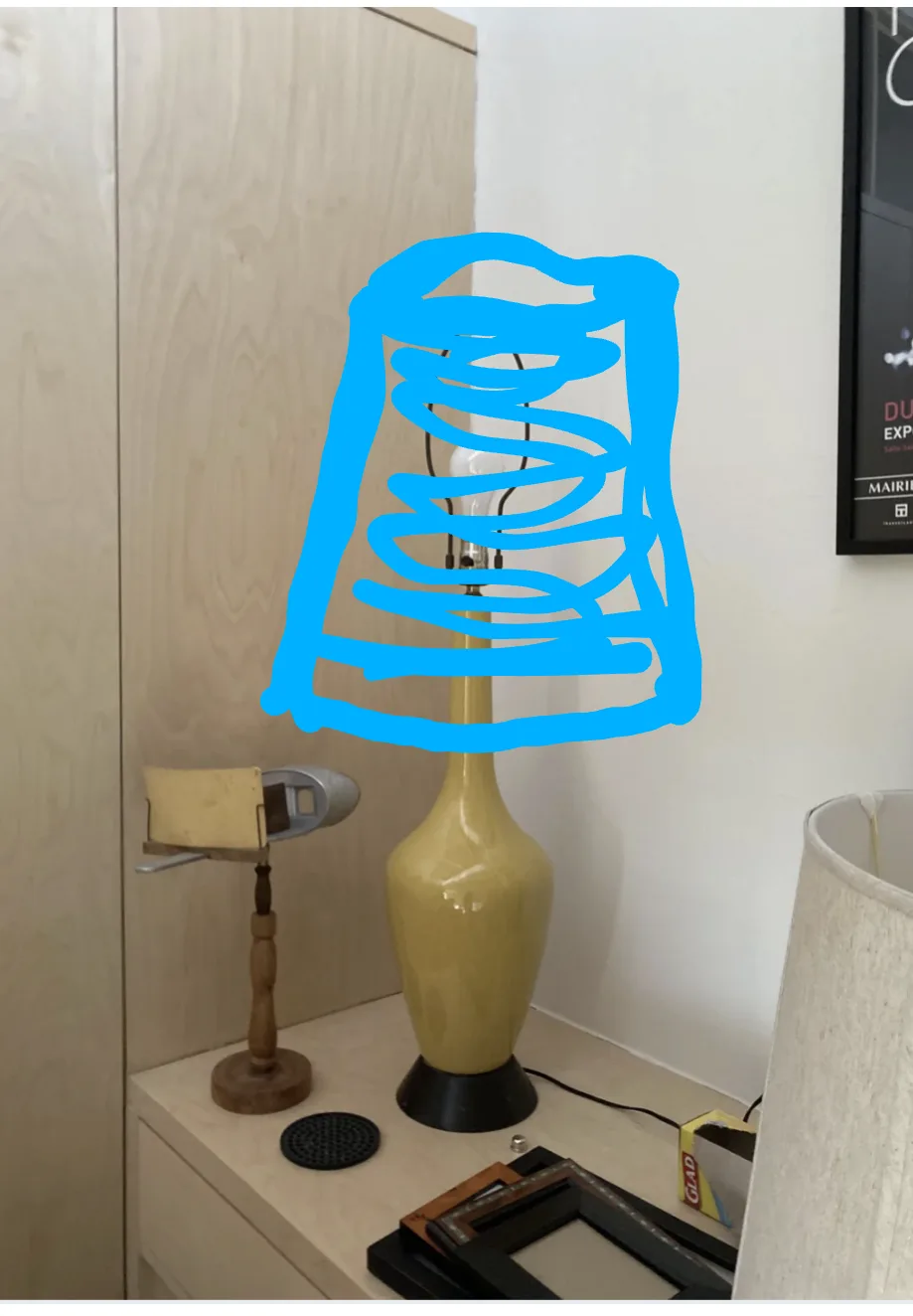 a yellow ceramic table lamp with a black base sits on a light-colored wooden dresser, its lampshade obscured by a large…