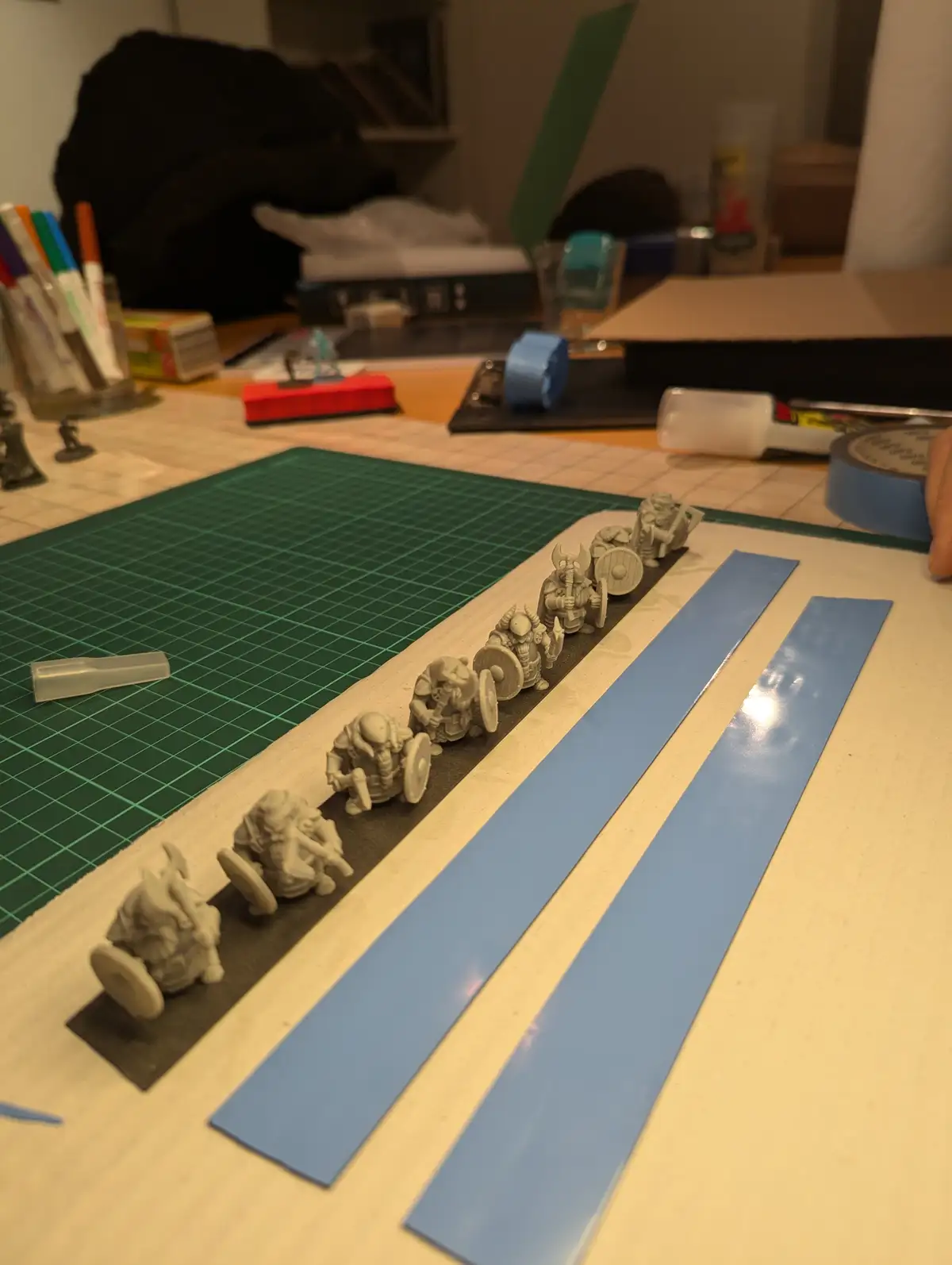 A row of unpainted fantasy miniatures, likely dwarves or similar warriors, are lined up on a black base on a worktable. Two…