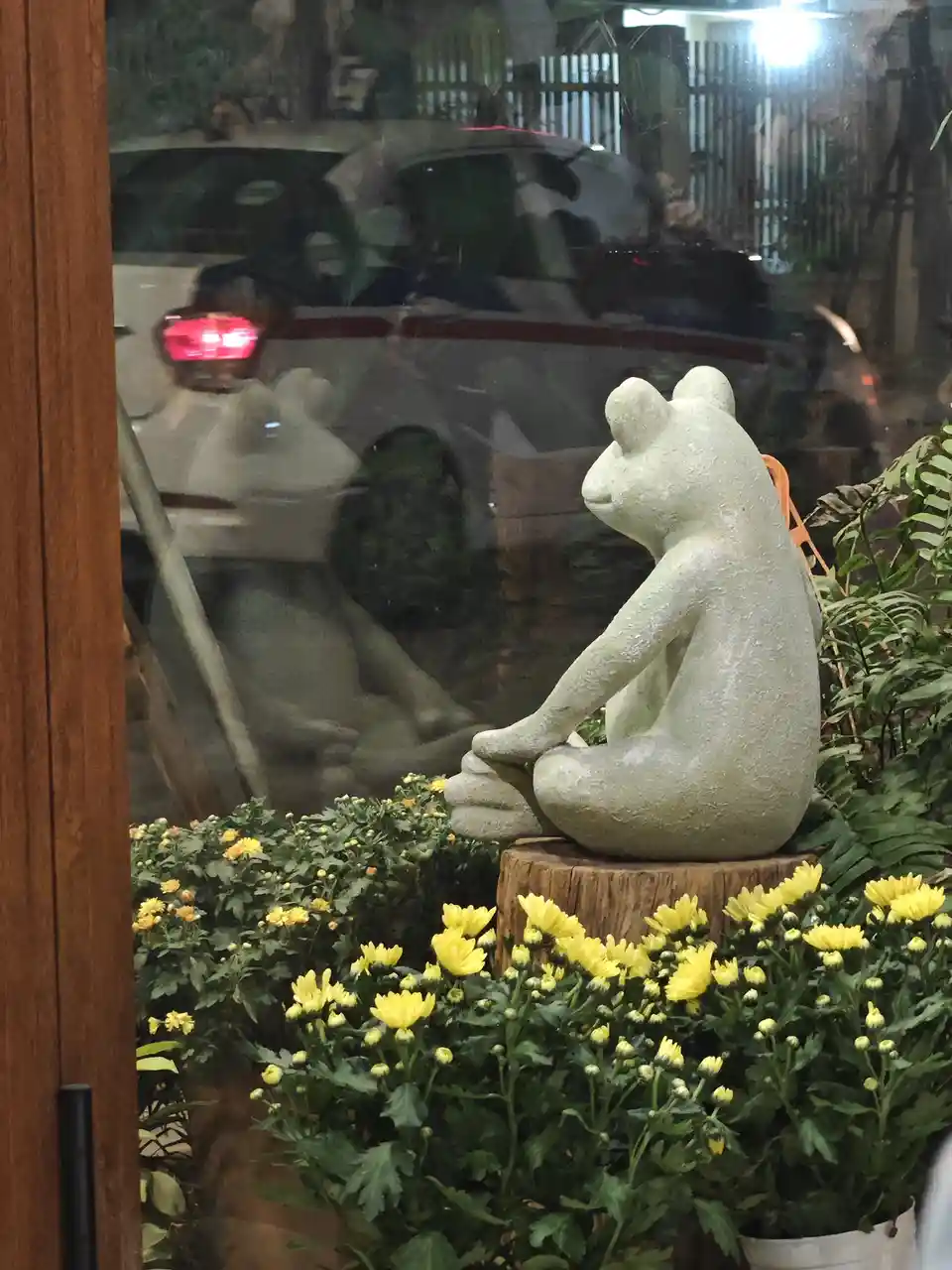 A stone frog statue sits cross-legged on a wooden stump surrounded by yellow flowers, viewed through a glass window…